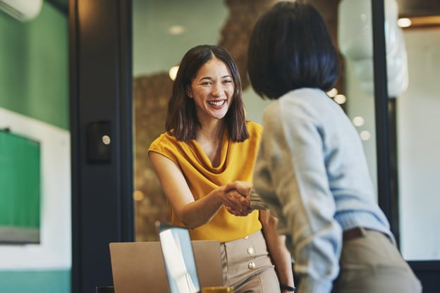 Two professional women shaking hands in a modern office, with one smiling at the camera.