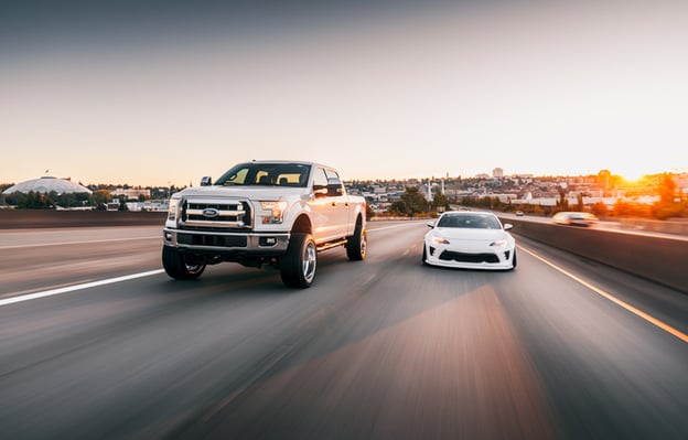 A white Ford pickup truck and a white sports car driving side by side on a highway at sunset.