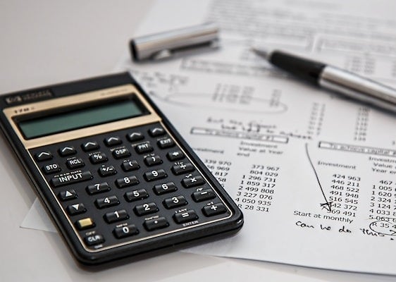 A financial calculator, a silver pen, and documents featuring investment tables and handwritten notes are arranged on a desk, illustrating a scene of financial planning or accounting.